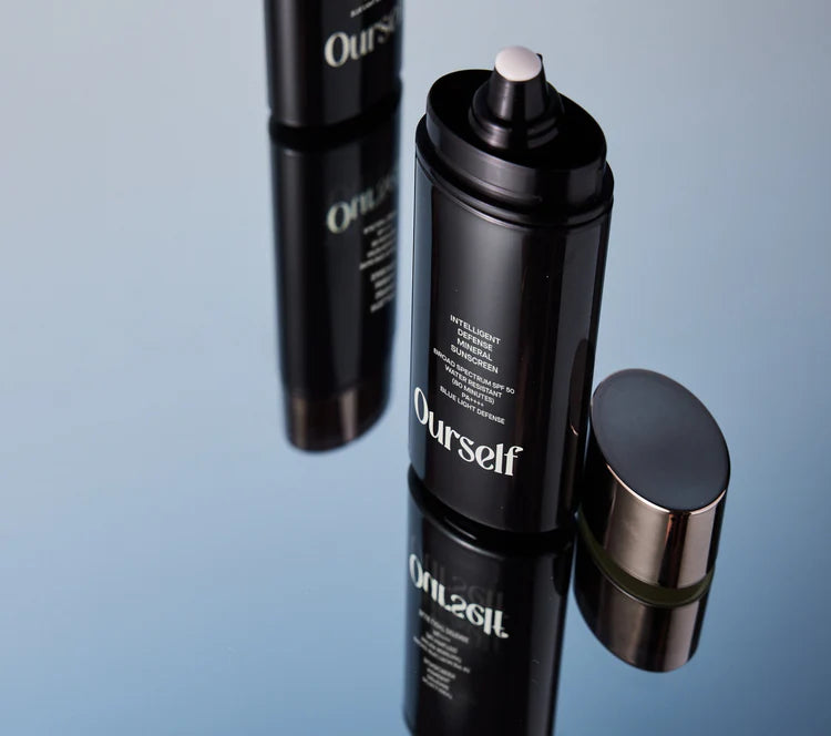 Black cosmetic bottles with 'Ourself Intelligent Defense Mineral Sunscreen Broad Spectrum SPF 50' branding on a reflective surface.